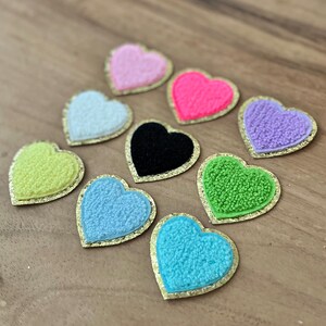 May include: A collection of colorful heart-shaped patches. Each patch features a textured, plush heart in various colors, including pink, black, green, and blue, with a gold-colored border. The patches are arranged on a wooden surface.