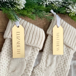 May include: Two white knitted stockings with gold gift tags. The tags are personalised with the names "Elizabeth" and "Matthew".