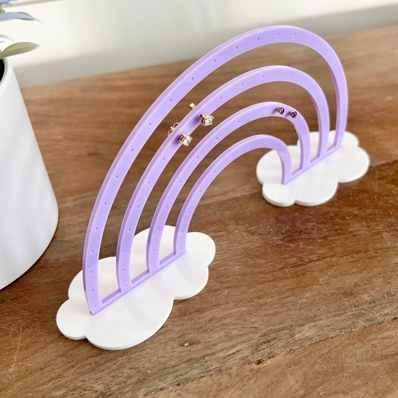 May include: A lavender rainbow-shaped earring holder with white cloud bases. The earring display has multiple holes for hanging earrings. Several pairs of earrings are displayed on the holder. The holder is on a wooden surface.