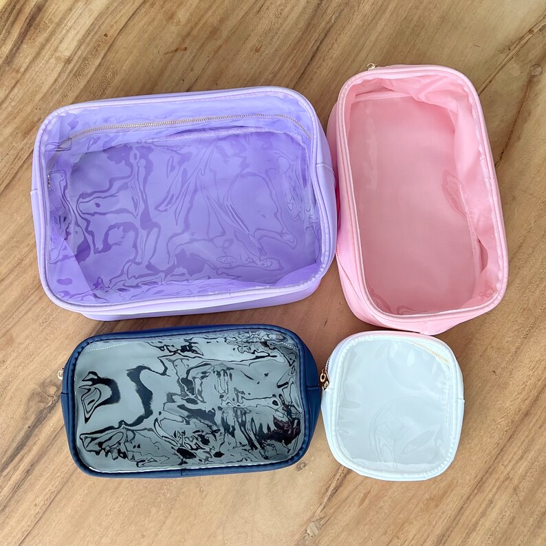 May include: Four clear makeup bags in different sizes and colors. The largest bag is lavender with a clear window, the second largest is pink, the third largest is navy blue with a clear window, and the smallest is white with a clear window.