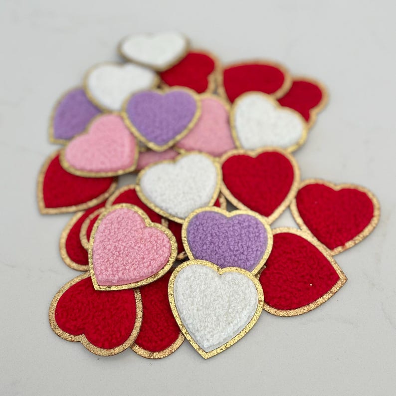 May include: A collection of heart-shaped patches in various colors, including red, white, pink, and purple. Each patch has a gold-colored border. The patches have a textured, fuzzy appearance.
