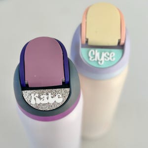 May include: Two personalized water bottles with colorful lids and names "Kate" and "Elyse" on the front. The bottles are white with a purple and gray band around the middle.