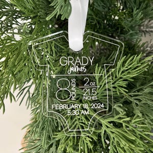 May include: Clear acrylic baby onesie ornament with the name "Grady James", birth weight of 8 pounds, 2 ounces, and length of 54.6 cm. The birth date is February 18, 2024, at 5:30 AM. A white ribbon is attached for hanging.