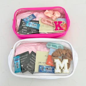 May include: Two clear cosmetic bags, one hot pink and one white, filled with various items. The pink bag contains a scrunchie, a letter "K" patch, and Orbit gum. The white bag includes a letter "M" patch, scrunchie, and makeup remover wipes.