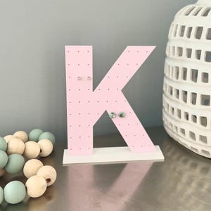 May include: A pink letter "K" shaped earring holder with small holes for displaying earrings. The holder is mounted on a white base. Several pairs of earrings are displayed on the holder. A white decorative vase is in the background.