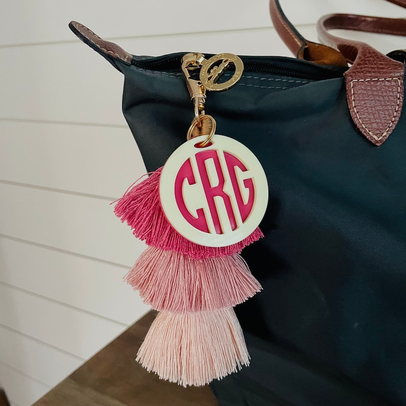 May include: A white circle keychain with a pink monogram "CRG" and a pink and white tassel. The keychain is attached to a black tote bag.