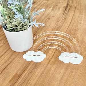 May include: Clear acrylic rainbow-shaped earring holder with white cloud bases. The earring holder has multiple curved arches and small holes for displaying earrings. The holder is on a wooden surface, with a white potted plant in the background.