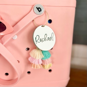May include: A white round tag with the name "Rachael" written in cursive script. The tag is attached to a pink tote bag with a gold ring and colorful tassels.