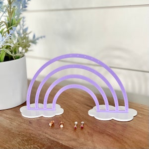 May include: A lavender acrylic earring holder shaped like a rainbow, with white cloud-shaped bases. The holder has multiple holes for displaying earrings. Several pairs of earrings are displayed in front of the holder. A white pot with blue flowers is in the background.