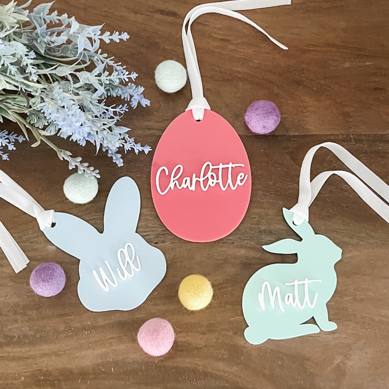 May include: Three Easter-themed gift tags with white ribbon. One tag is a pink egg with the name "Charlotte" written on it. Another tag is a light blue bunny with the name "Will" written on it. The third tag is a light green bunny with the name "Matt" written on it.
