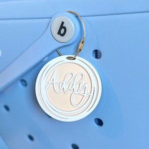 May include: A white personalized name tag with the name "Addy" written in a cursive font. The tag is attached to a blue tote bag with a gold metal hook.
