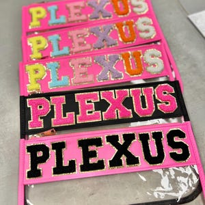 May include: A collection of pink and black zippered pouches, each displaying the word "PLEXUS" in vibrant, textured lettering. The pouches feature a clear front and a gold-toned zip. Lettering colours include yellow, blue, pink, purple, and orange.