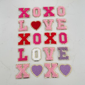 May include: A collection of embroidered patches in various colors, including pink, red, and purple. The patches feature the words "XOXO" and "LOVE", along with heart shapes. Each patch has a gold border.