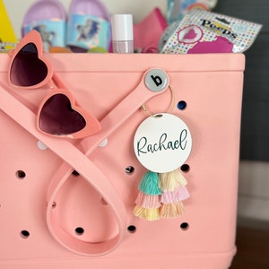 May include: A pink tote bag with a white circle tag that says "Rachael" hanging from the handle. The tag has a colorful tassel attached to it.