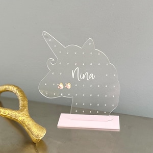 May include: Clear acrylic unicorn-shaped earring holder with the name "Nina" in cursive script. The holder has multiple small holes for earrings and sits on a pink base. Two small unicorn earrings are displayed.