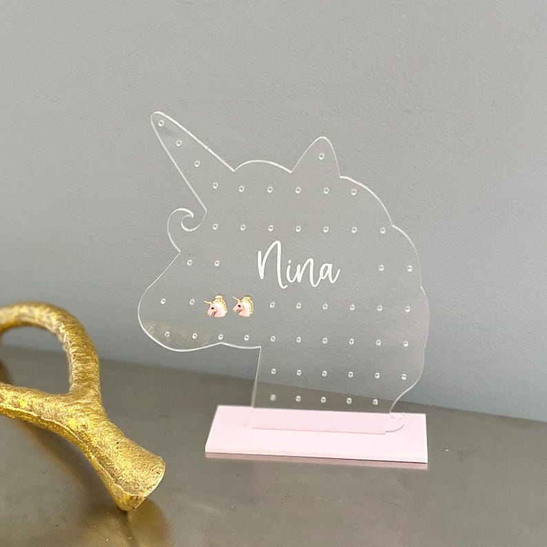 May include: Clear acrylic unicorn-shaped earring holder with the name "Nina" in cursive script. The holder has multiple small holes for earrings and sits on a pink base. Two small unicorn earrings are displayed.