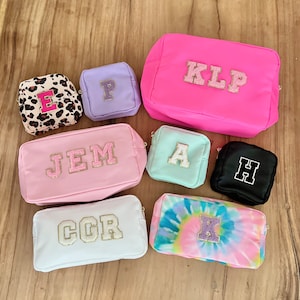 Peut inclure: Assortiment de huit trousses de toilette roses, blanches, noires, violettes et bleues avec des lettres dorées pailletées épellant les noms "JEM", "KLP", "CCR", "E", "P", "A", "H" et "K".