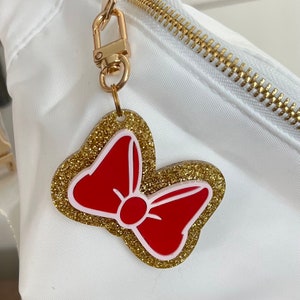 May include: A gold glitter bow charm with a red bow in the center. The charm is attached to a gold key ring.