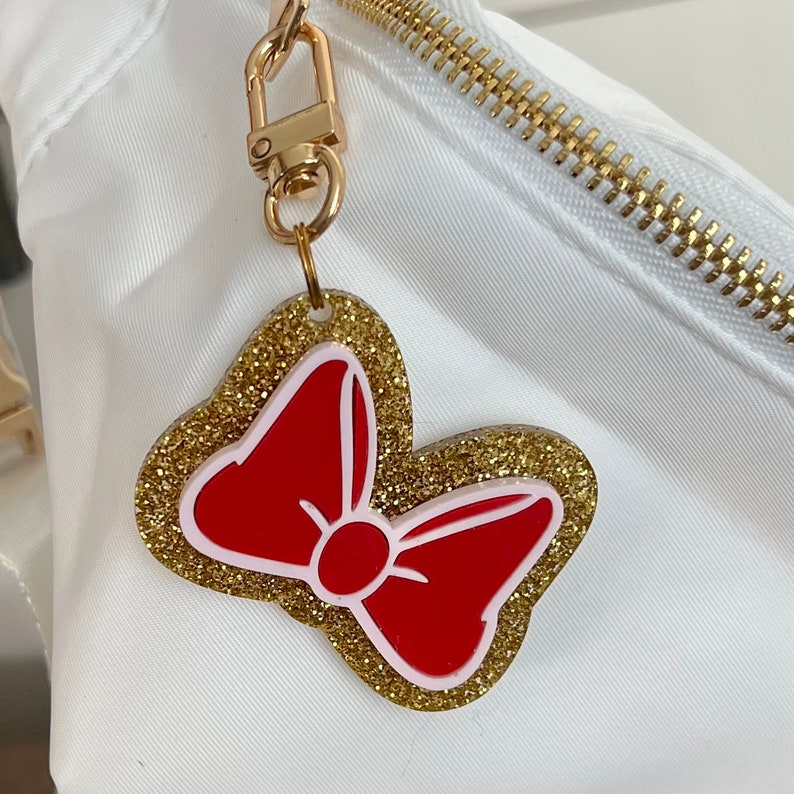 May include: A gold glitter bow charm with a red bow in the center. The charm is attached to a gold key ring.