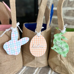 May include: Three burlap Easter baskets with bunny ears and personalized tags. The tags are shaped like an egg and a bunny and are decorated with a gingham pattern. The tags are personalized with the names "Mom", "Andrew", and "Thomas".