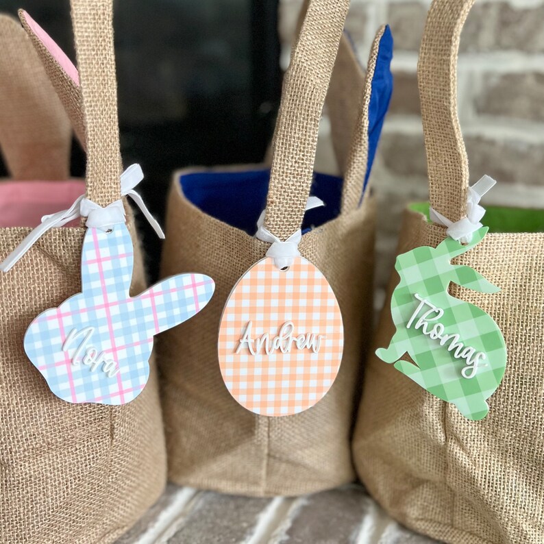 May include: Three burlap Easter baskets with bunny ears and personalized tags. The tags are shaped like an egg and a bunny and are decorated with a gingham pattern. The tags are personalized with the names "Mom", "Andrew", and "Thomas".