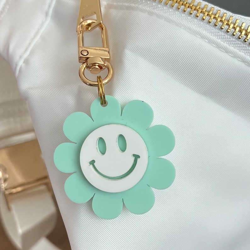 May include: A light green flower-shaped charm with a white smiley face. The charm is attached to a gold chain.