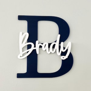 May include: A navy blue wooden letter "B" with the name "Brady" in white script layered on top. The personalized wall decor is a decorative item for a nursery or child's room.