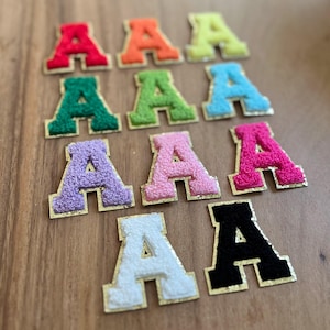 May include: A selection of embroidered letter "A" patches in a variety of colours. The patches have a fuzzy texture and are in shades of red, orange, yellow, green, blue, purple, pink, white, and black, with gold-coloured borders.
