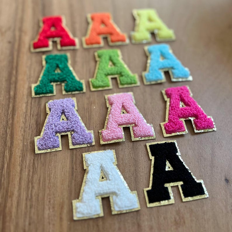 May include: A selection of embroidered letter "A" patches in a variety of colours. The patches have a fuzzy texture and are in shades of red, orange, yellow, green, blue, purple, pink, white, and black, with gold-coloured borders.