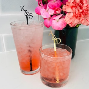 May include: Two glasses of pink cocktails with ice, each featuring a personalized drink stirrer. One stirrer is black with the letters "K&S", the other is gold with "A&D". A vase of pink flowers is in the background.