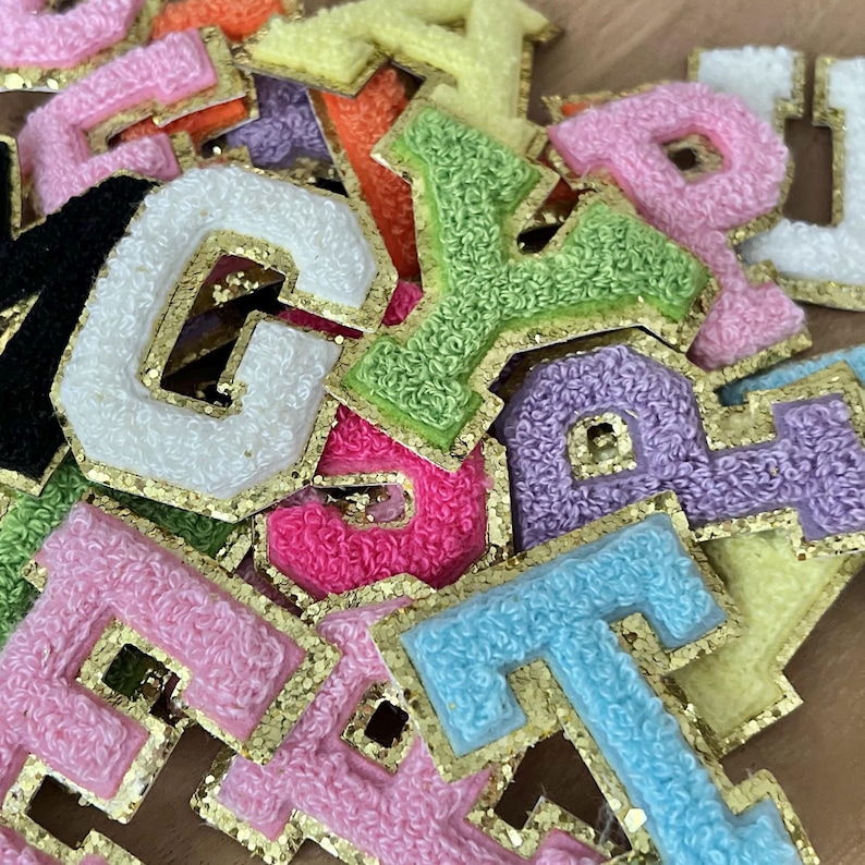 May include: A selection of colourful, embroidered fabric letters in shades like pink, green, and blue. Each letter is edged with gold glitter and has a textured, fuzzy look. The letters are scattered, creating a vibrant and playful display.