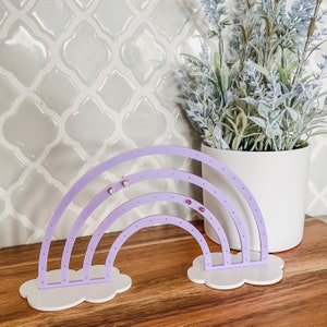 May include: A lavender rainbow-shaped earring holder with white cloud-shaped bases. The earring holder has multiple holes for displaying earrings. The background includes a white pot with faux lavender and a light gray tile wall.