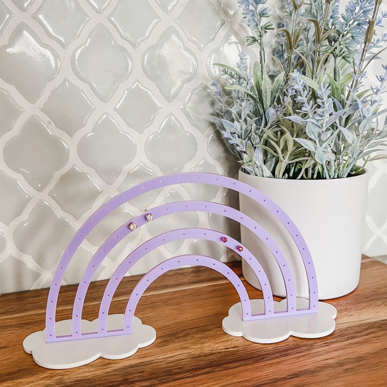 May include: A lavender rainbow-shaped earring holder with white cloud-shaped bases. The earring holder has multiple holes for displaying earrings. The background includes a white pot with faux lavender and a light gray tile wall.