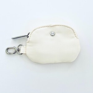 Puede incluir: Una peque&ntilde;a bolsa de cremallera de color blanco roto con un tirador de cremallera plateado y un cierre de presi&oacute;n plateado. La bolsa tiene un peque&ntilde;o logotipo plateado en la parte delantera.