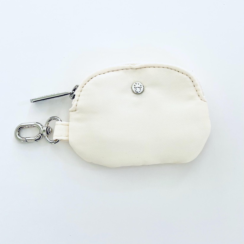 Puede incluir: Una peque&ntilde;a bolsa de cremallera de color blanco roto con un tirador de cremallera plateado y un cierre de presi&oacute;n plateado. La bolsa tiene un peque&ntilde;o logotipo plateado en la parte delantera.