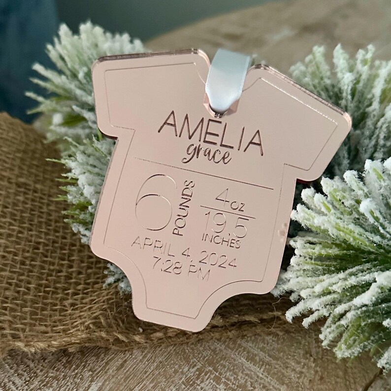 May include: A rose gold baby onesie-shaped ornament with the name "Amelia Grace" engraved on it. The ornament also includes the baby's weight (2.72 kg, 113.4 grams), length (49.5 cm), and birth date and time: April 4, 2024, 7:28 PM.