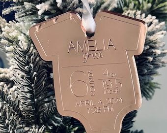 May include: A rose gold baby onesie-shaped ornament with the name "Amelia Grace" engraved on it. The ornament also includes the baby's weight, length, and birth date: 6 pounds, 4 ounces, 19.5 inches, April 4, 2024, 7:28 PM. A white ribbon is attached.
