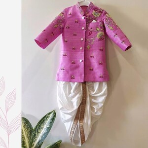 May include: A children's formal outfit. It features a bright pink jacket with detailed embroidery and white buttons. The jacket is paired with white dhoti trousers, creating a traditional look. The jacket has a pocket on the left side.