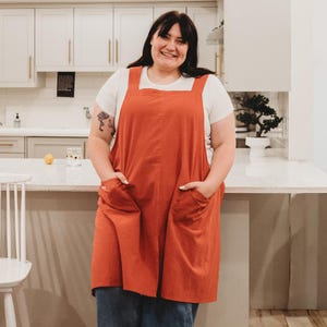 May include: A solid burnt orange apron featuring wide shoulder straps and two large front patch pockets. The apron is worn over a plain white short-sleeved t-shirt and blue denim jeans, creating a functional and simple layered look for daily tasks.