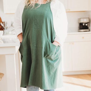 May include: A solid forest green apron featuring a square neckline, thick shoulder straps, and two large patch pockets on the front. The apron is worn over a white long-sleeved shirt and blue jeans, providing full coverage for the torso and upper legs.