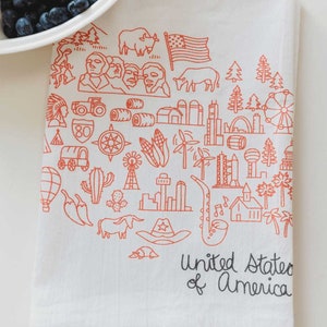May include: A white tea towel with an orange outline of various icons representing the United States of America. The icons include a cowboy hat, a cactus, a hot air balloon, a tractor, a compass, a windmill, a skyscraper, a saxophone, a corn cob, a bald eagle, a mountain range, a flag, a house, a palm tree, a musical note, and a wagon. The text "United States of America" is written in black at the bottom of the towel.