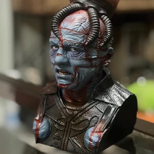 May include: A detailed bust of a character with blue skin, adorned with a network of wires and a bulbous headpiece. The figure has a stern expression, with red accents suggesting blood. The bust features a dark, textured armor-like chest piece.
