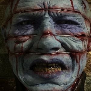 May include: A close-up of a blue and red horror mask with stitched-up eyes and mouth. The mask is covered in blood and has a distressed look.