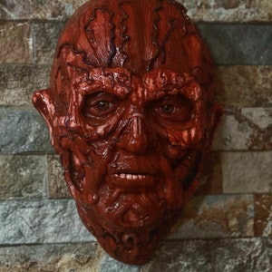 May include: A red and white horror mask with a detailed, textured surface. The mask depicts a human face with a grotesque, melted appearance.