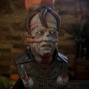 May include: A detailed bust of a pale-skinned figure with a large orange headpiece and dark, spiraling horns. The figure has blood-like streaks across the face and chest, with a stern expression. The bust is set against a blurred background.