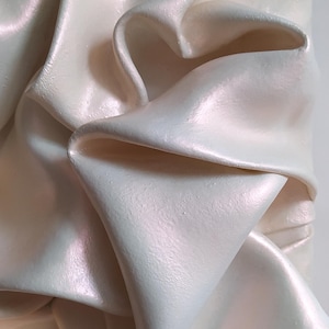 May include: Close-up of a sculpted, off-white, pearlescent object with a rippled, fabric-like texture. The folds and curves create shadows, highlighting the object's three-dimensional form. The surface has a subtle sheen, suggesting a smooth finish.