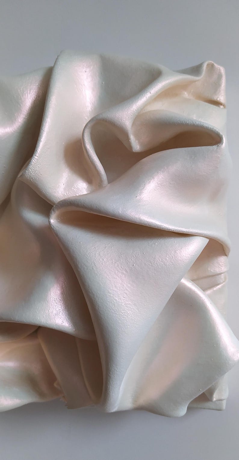May include: Close-up of a sculpted, off-white, pearlescent object with a rippled, fabric-like texture. The folds and curves create shadows, highlighting the object's three-dimensional form. The surface has a subtle sheen, suggesting a smooth finish.