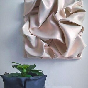 May include: A square, cream-colored wall art piece with a textured, draped fabric design. Below, a dark blue square planter holds a small green plant. The art and plant sit against a white wall and table.