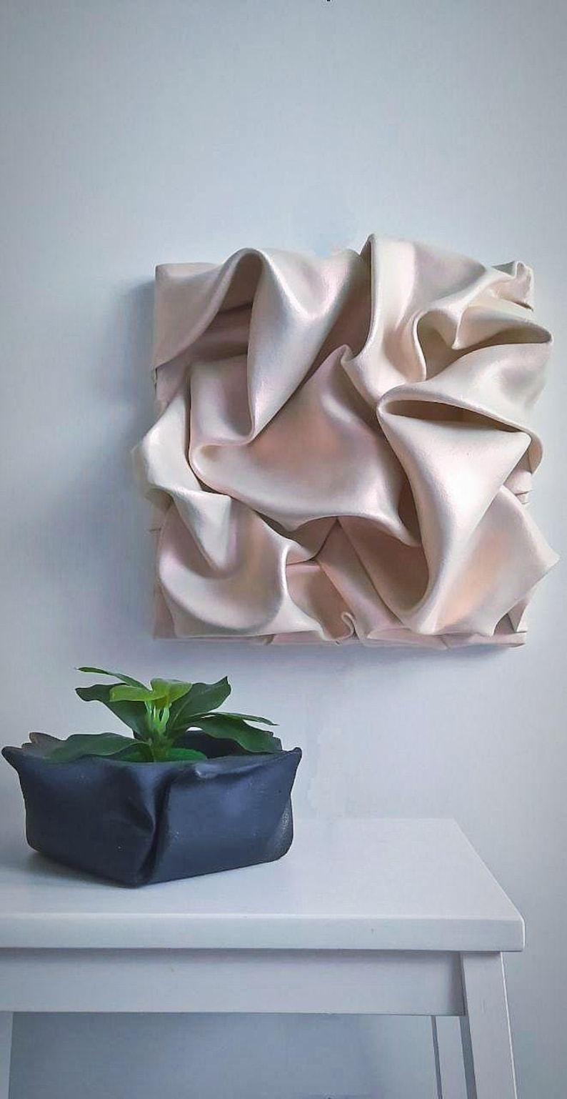 May include: A square, cream-colored wall art piece with a textured, draped fabric design. Below, a dark blue square planter holds a small green plant. The art and plant sit against a white wall and table.