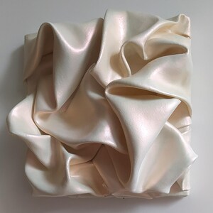 May include: A square, off-white sculpture with a rippled, fabric-like texture. The surface has a subtle pearlescent sheen, catching the light and creating highlights and shadows. The artwork is mounted on a white background.